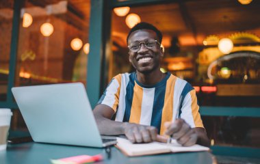 Portrait of happy dark skinned male copywriter with education notebook working remotely in street cafe, successful hipster blogger in optical eyeglasses smiling at camera during laptop e learning