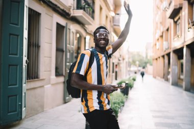 Cheerful young black guy in striped tshirt with backpack turning back saying goodbye with raised hand while stroll on narrow street of old buildings in summer day