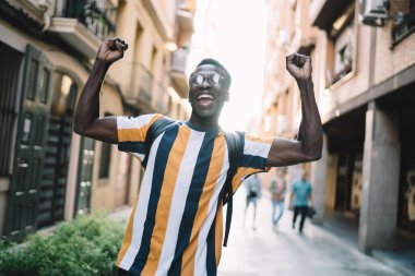 Excited male traveller dressed in trendy outfit raising hands rejoicing in city and feeling satisfied with touristic journey for recreating, joyful hipster guy in classic eyewear having fun