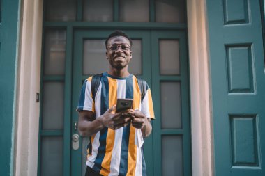 Half length portrait of cheerful dark skinned blogger holding smartphone device in hands and smiling at camera, happy African American tourist with cellphone enjoying solo vacations for visiting city