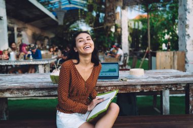 Optimistic Asian female freelancer in casual clothes smiling and looking at camera while holding notebook in hands during remote work