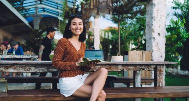 Positive Asian female freelancer in casual clothing smiling and looking at camera while sitting on wooden bench and writing ideas