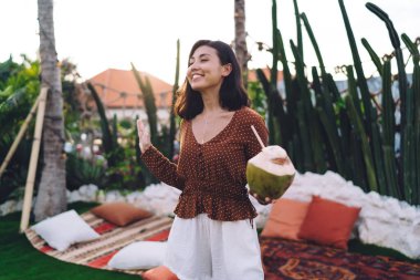 Young ethnic brunette in brown polka dot blouse and white shorts enjoying fresh coconut water and smiling happily with eyes closed while standing on open terrace of resort house