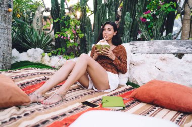 Full body of happy ethnic woman in casual clothes sitting on comfortable soft blanket in tropical garden and drinking cocktail while relaxing and looking at camera