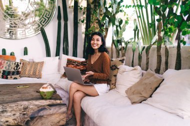 Optimistic female in casual clothes sitting on comfortable sofa and using laptop while resting in living room and smiling at camera