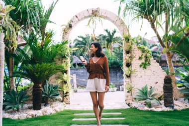Content female tourist in summer clothes with mobile phone in hand walking in courtyard near green hedge and palms and looking away