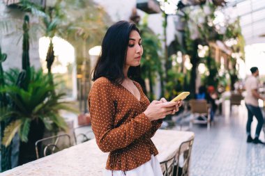 Side view of serious female manager in stylish outfit leaning on table while typing message about project and working remotely from modern cafe
