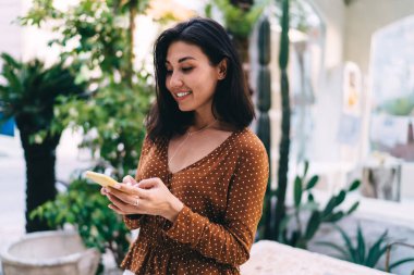 Side view of optimistic female in trendy clothes answering messages on smartphone and standing near green potted bushes in city cafe