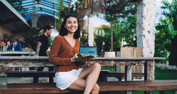 身穿休闲装的积极的亚洲女自由职业者坐在木制长椅上 一边微笑着 一边看着相机 一边写着自己的想法 — 图库照片