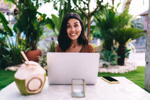 身着休闲装的年轻亚洲女性坐在笔记本电脑前 坐在露天咖啡店的桌子前 一边看着相机 一边用智能手机和椰子喝水 这让她们震惊极了 — 图库照片
