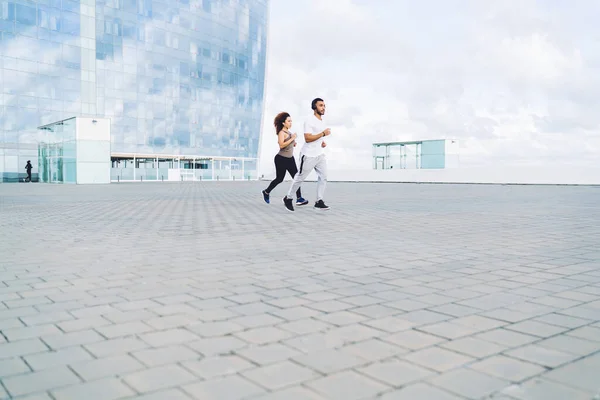 運動中に近代的なガラスの建物の近くの舗装上のスポーツウェアジョギングで若い民族の男性と女性の完全な体外 — ストック写真