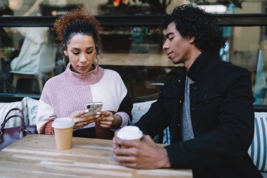 Farklı siyah ceketli genç adam modern kafede kahve içerken cep telefonu kullanan gündelik kazaklı bayana bakıyor.