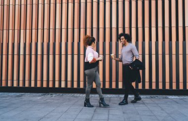 Pozitif Afro-Amerikan çiftin gündelik kıyafetleri içinde modern binanın yanında durup kahve içerken birbirlerine bakmaları.