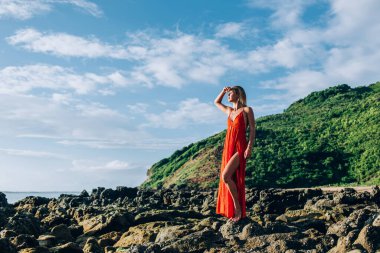 Yeşil tepe adasının huzurlu sahil kıyısında dururken, parlak kırmızı elbisesiyle gözlerini kapayan ve düşüncelere dalan düşünceli kadının yan görüntüsü
