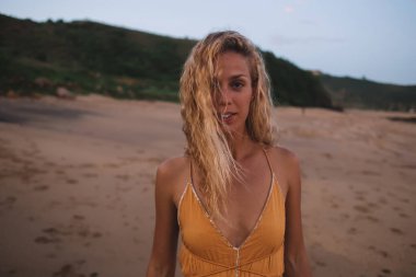 Dertsiz, renkli elbiseli güzel kadın kameraya bakıyor ve rüzgarlı bir günde çimenli tepenin kenarındaki tropikal sahilde geziniyor.