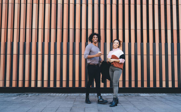 Full length of positive African American couple in casual clothes standing near modern building and drinking coffee while talking and looking at camera