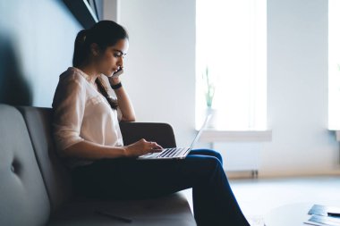Ofiste cep telefonuyla konuşurken dizüstü bilgisayarlı klavyede yazan resmi giyinen odaklı etnik kadın yan görünümü