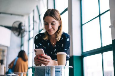 Bluzlu ve kot pantolonlu gülümseyen genç bayan güneş ışığında pencerenin yanında akıllı telefon kullanırken bir fincan kahveyle masaya yaslanıyor.
