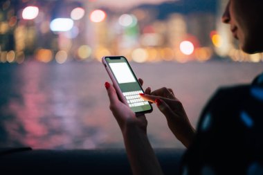 Tanımlanamayan bir kadın, pembe çantalı modern akıllı telefon kullanıyor. Ulak, gece nehri ve Bokeh şehir ışıklarının bulanık arka planında maket yapıyor.