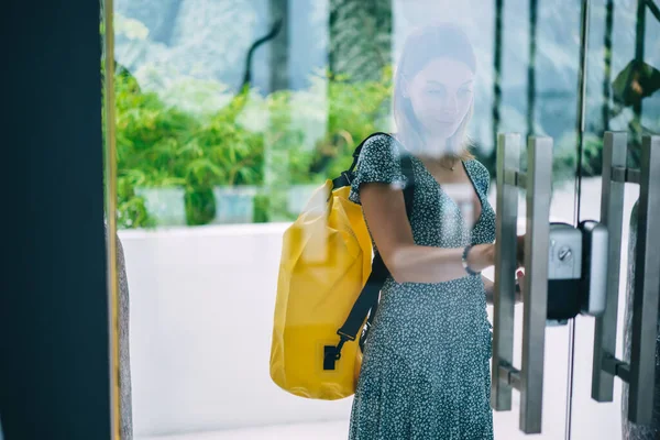 若いですティーンCaucasian女性で流行のカジュアルドレスで黄色のバックパックが入る建物 魅力的なヒップスター女の子でリュックサック付きガラスドアを引っ張っ反射で外部からセキュリティキーを使用して — ストック写真