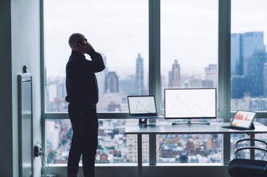 Resmi giyinmiş, gözlüklü, modern ofiste duran ve cep telefonuyla konuşan yüzü olmayan erkek çalışanın yan görüntüsü.