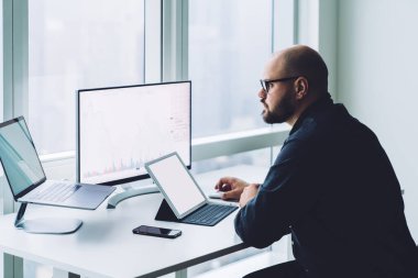 Bilgisayarda internette gezinen düşünceli bir adamın yan görünüşü. Elinde aletlerle oturmuş, dünya hisselerinde bitcoin ticareti yapıyor.