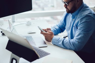 İş sırasında akıllı telefondan mesajlaşan resmi giyimli ve gözlüklü, konsantre sakallı erkek girişimcinin tepesinden.