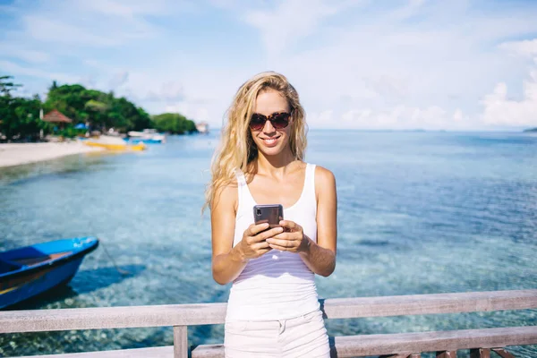 社会的なフォロワーとスマートフォンのメッセージを楽しむサングラスの陽気な女性はモルディブの休暇について通信し 笑顔の女性観光客が旅行ブログを共有するためのテキスト出版物を入力 — ストック写真