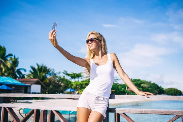 インドネシアでの休暇中にセルフィーメディアコンテンツを作るために携帯電話のフロントカメラを使用してサングラスで幸せな白人観光客 陽気な若い女性撮影の影響ビデオと笑顔 — ストック写真