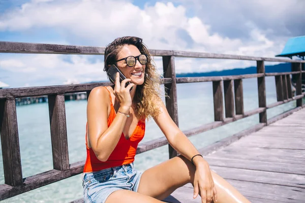 穿着太阳镜和泳衣的无忧无虑的晒黑女人坐在木制码头上 一边用手机聊天 一边在海滨大笑 — 图库照片