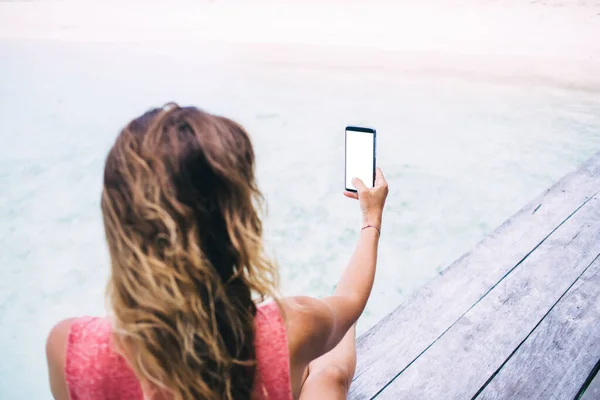 現代のデジタル装置で撮影した巻き毛の女性のバックビューと海の近くの晴れた日に木製の桟橋に座って — ストック写真