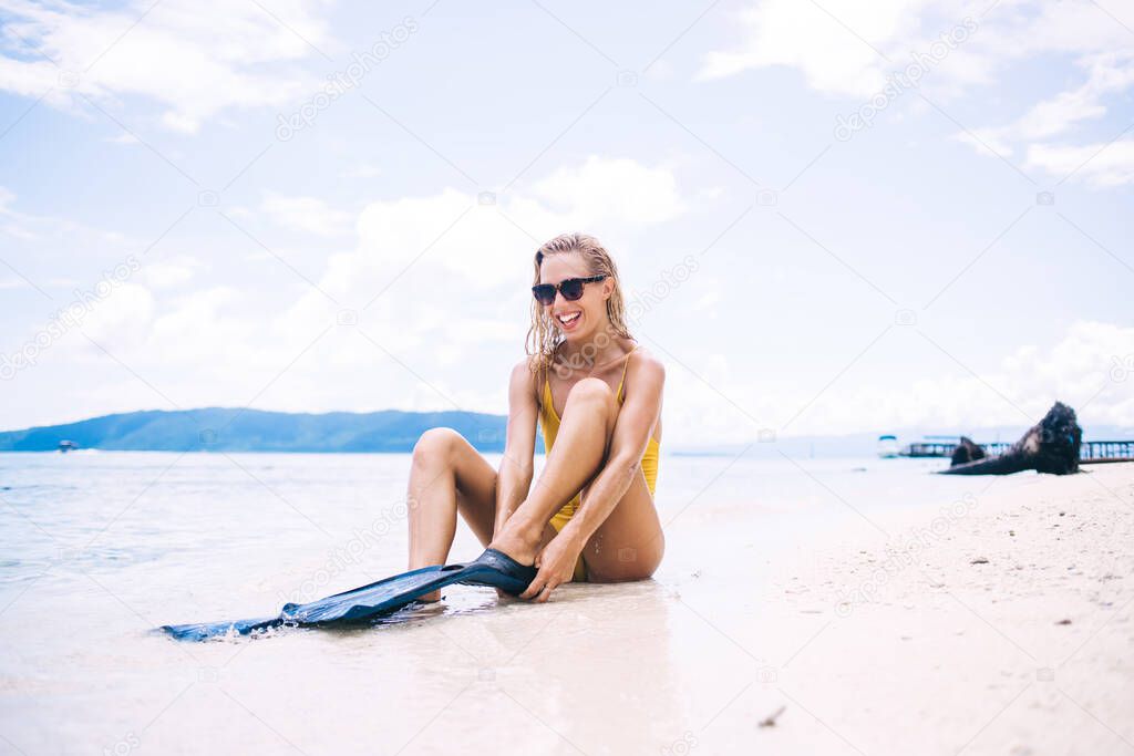 Joyful Caucasian swimmer in sunglasses sitting at sea coastline and wearing flippers for practicing hobby in water, happy woman with swim fins laughing at tropical beach satisfied with getaway trip