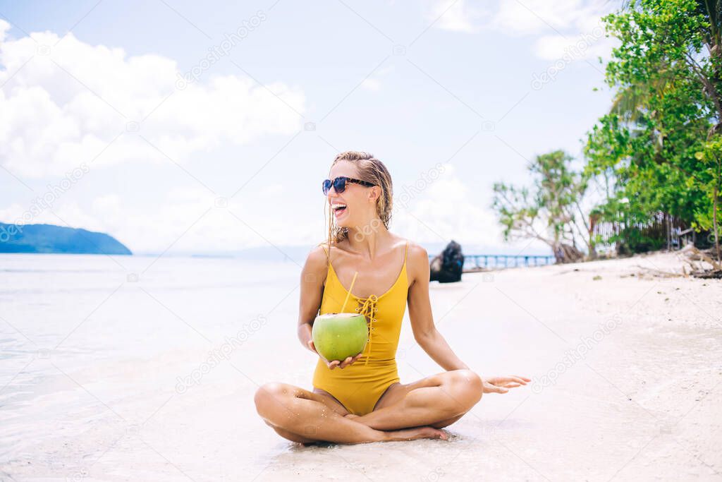 Happy Caucasian woman with exotic fruit sitting at white sand of Maldives and smiling, pretty female swimmer holding coconut beverage enjoying free time for sunbathing on Bahamas coastline