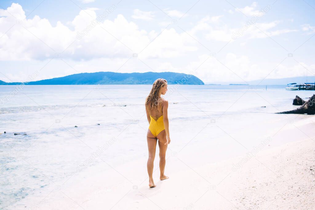 Slim Caucasian woman in yellow swimsuit walking at sandy beach of Mauritius island exploring tropical nature environment during sunbathing, charming female tourist recreating in resort paradise