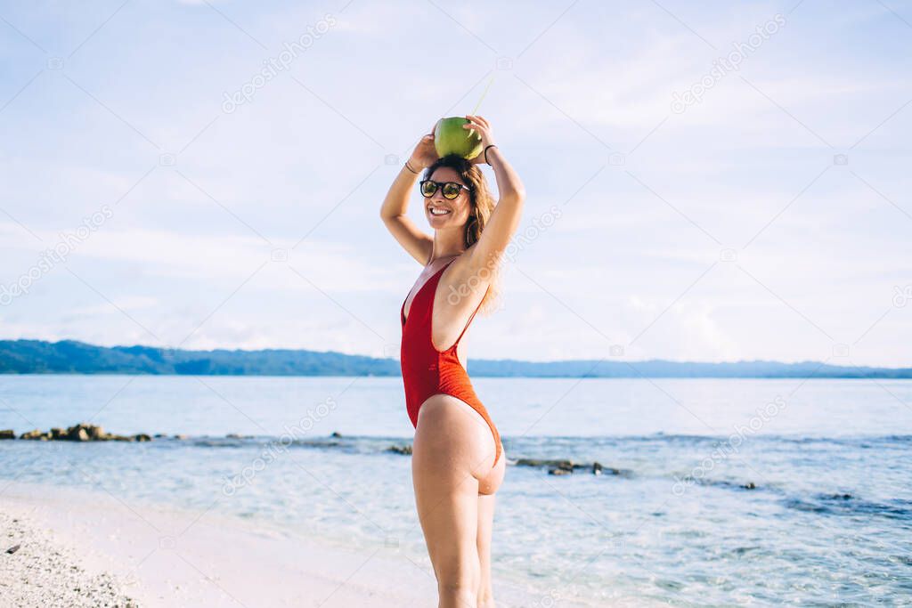 Half length portrait of cheerful female with coconut fruit on head smiling at camera enjoying perfect vacations on Maldive, happy woman in sunglasses spending time for sunbathing on tropical beach