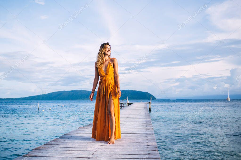 Carefree woman in trendy sundress standing at wooden pier enjoying vacations time on Raja Ampat island, happy woman smiling during resort travel journey for visiting tropical nature environment