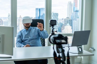 VR gözlüklü bir erkek, modern ofis manzaralı pencereli bilgisayarlarla masada otururken çekimlere katılıyor.