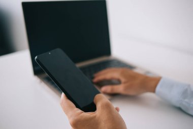 Yukarıdan gelen bilgilere göre, isimsiz bir girişimci elinde dizüstü bilgisayarla ahşap masada oturuyor ve evde uzaktan çalışırken akıllı telefonlara bakıyor.