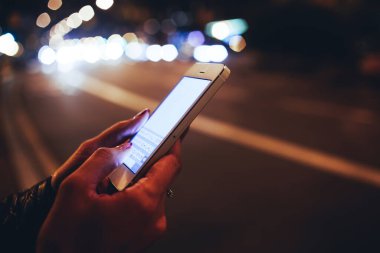 Siyah deri ceketli, arka plandaki cep telefonu ekranını gösteren kimliği belirsiz bir kadın gece atmosferik şehirde ışık saçar.