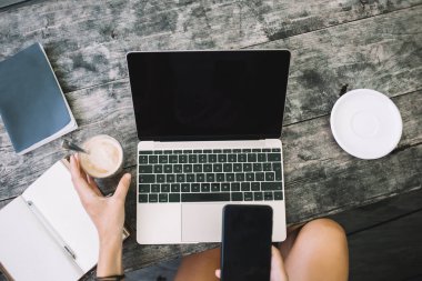 Akıllı telefon kullanan ve kahve içen, modern dizüstü bilgisayar ve dizüstü bilgisayarlı ahşap masada oturan...