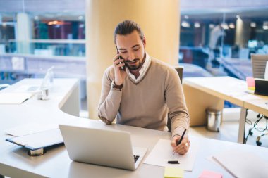 Gündelik kıyafetlerle gezen, cep telefonuyla iş stratejisini tartışan ve modern iş yerindeki dizüstü bilgisayarla masada otururken kağıda not alan erkek girişimci.