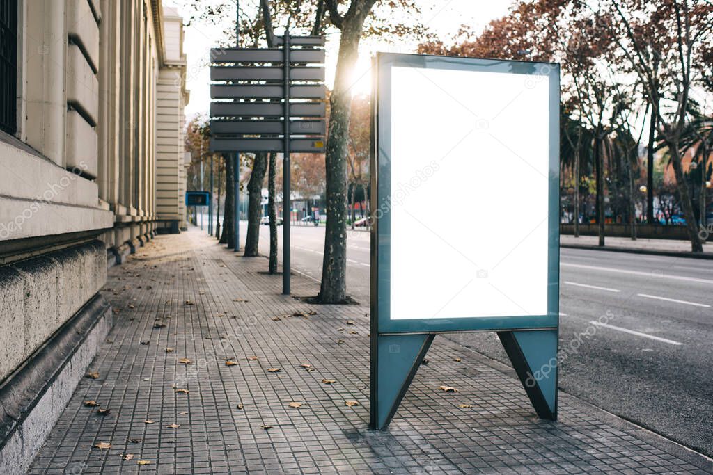 Empty white advertising vertical placard for promotional content placed in city street near empty road and buildings on autumn day