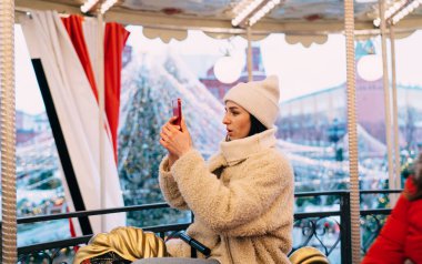 Sıcak giysili, şapkalı, atlıkarınca süren ve akıllı telefondan fotoğraf çeken heyecanlı bir kadın.