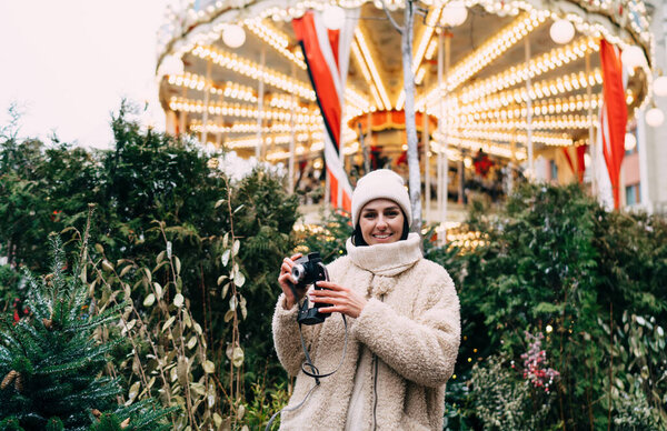 Low angle of happy woman in warm wear and hat smiling and taking photo on vintage photo camera while looking at camera