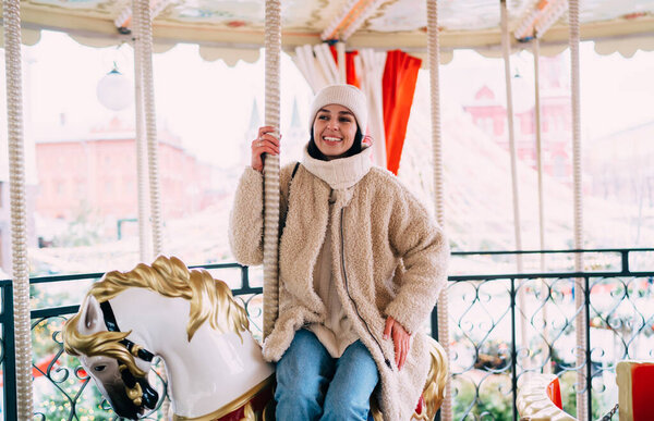 Happy female in warm outerwear and hat smiling and looking away while riding horse on carousel in Christmas fairground in evening