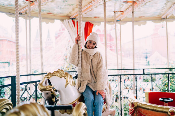 Happy smiling female in warm clothing and hat looking at camera while sitting on horse on illuminated Christmas carousel in park