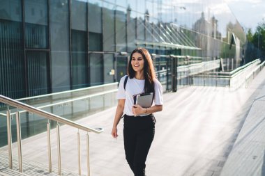 Modern dijital tabletli mutlu Kafkasyalı öğrenci şehir merkezindeki üniversite binasının yanında yürüyor, tasasız hippi kız dokunmatik ped tutuyor ve açık havada gülümsüyor.