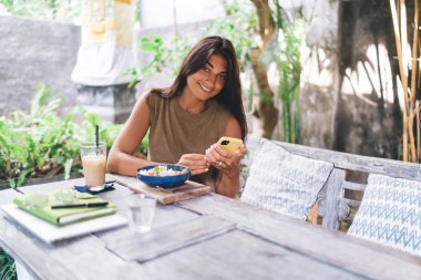 Açık hava restoranında yemek masasında oturup kameraya bakarken günlük kıyafetleriyle gülümseyen ve akıllı telefonlara bakan genç etnik bir kadın.