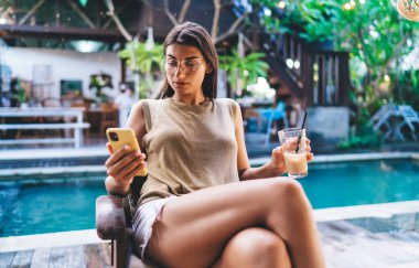 Gözlüklü, elinde bir bardak içecek ve cep telefonuyla yüzme havuzunun yanındaki sandalyede oturan odaklanmış etnik kadın.