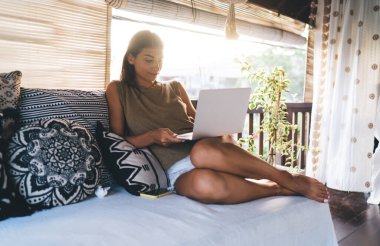 Çıplak ayaklı, rahat bir odada rahat bir koltukta dinlenen, arka koltukta dizüstü bilgisayar kullanan, gündelik kıyafetleriyle etnik bir kadın.